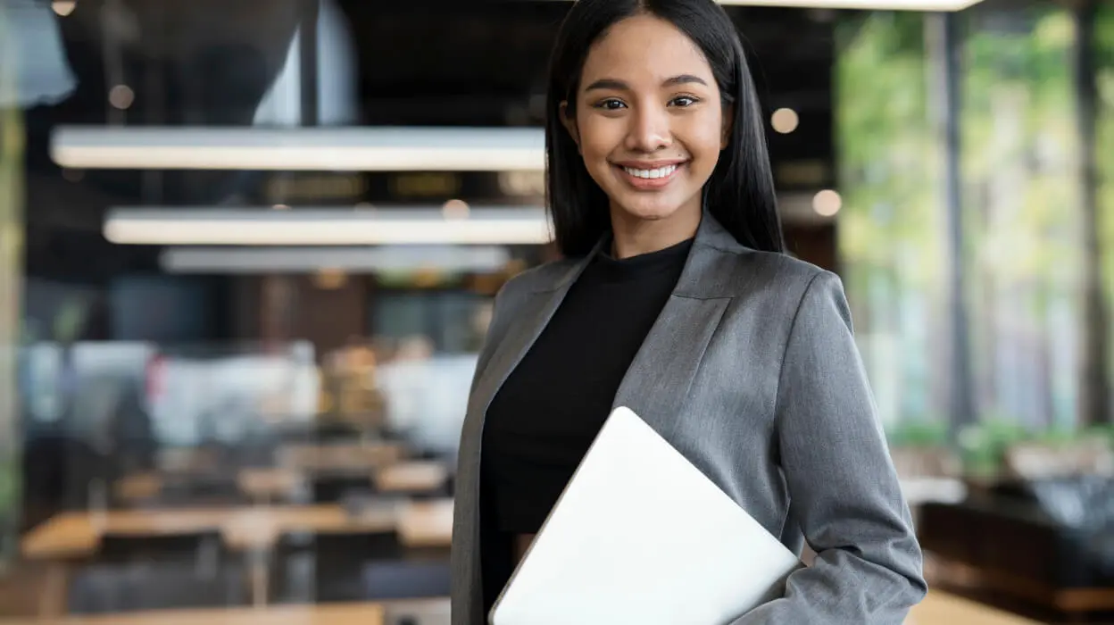 A cheerful young woman is holding a laptop, showcasing a bright smile and a positive demeanor.