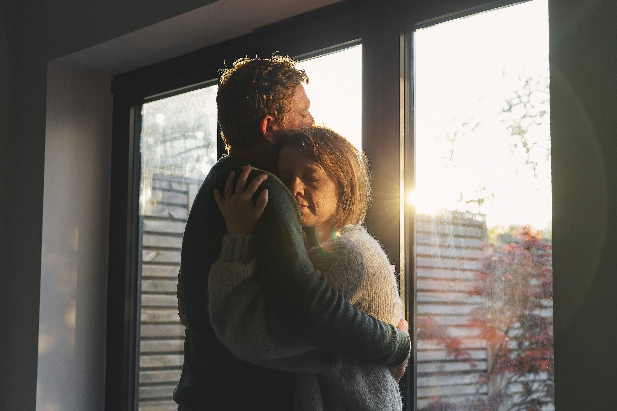 Mature couple embrace while standing near a window with sun streaming in